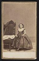 Photograph:  M. Lavinia Warren standing beside a chair, both hands on her dress